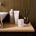 NEOM Super Shower Power Body Cleanser, Magnesium Body Butter, scented candle and reed diffuser styled together on a wooden bathroom shelf.