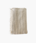 A folded beige throw blanket with thin black stripes and fringed edges. The fabric appears textured and soft, evoking a sense of warmth and comfort. | SW Home